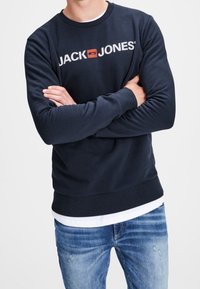 Un uomo indossa un felpa navy blue di Jack & Jones con le braccia incrociate, abbinata a jeans blu sbiaditi su uno sfondo bianco semplice.