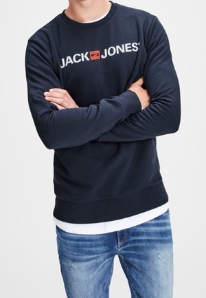 Un uomo indossa un felpa navy blue di Jack & Jones con le braccia incrociate, abbinata a jeans blu sbiaditi su uno sfondo bianco semplice.