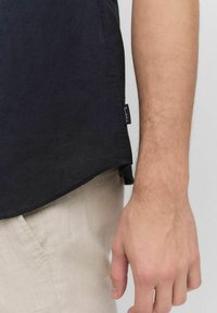 Mörkblå skjorta med rundad nederkant, tillverkad av mjukt tyg. Har en liten etikett på vänster sida. Ljust färgade, strukturerade byxor synliga.