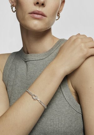 Bracciale in argento con chiusura a toggle, indossato su un braccio di carnagione chiara. Lo sfondo presenta una maglietta senza maniche grigia e texturizzata.