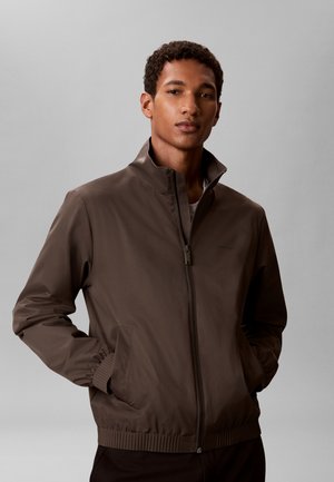 Joven con chaqueta marrón con cremallera y las manos en los bolsillos, de pie frente a un fondo gris liso.