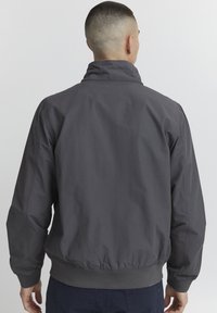 Jachetă bomber gri cu guler independent, manșete și tiv elastic, dintr-un material neted și ușor. Cusături întărite vizibile.
