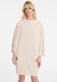 Beiges Kleid aus glattem Stoff, mit lockerer Silhouette, Fledermausärmeln und zwei vorderen Taschen mit dekorativen Akzenten.