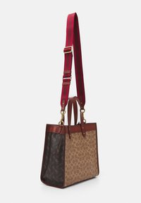 Brun og tan tote taske med et totonet design, der har et brunt mønstret stof og tan monogramtrykt, akcentueret med brune læderkanter og en rødstribet rem.