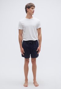 Άσπρο βαμβακερό t-shirt και σορτς με ναυτικό ρίγες, που διαθέτουν ελαστική μέση και κορδόνι. Το μοντέλο στέκεται ξυπόλυτο μπροστά σε απλό φόντο.