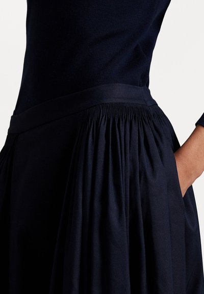 Sieviete, ģērbusies tumši zilos pieguļošos topā un plisētā svārkos ar plašu jostu, roka nevainojami ielikta svārku kabatā.