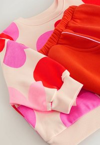 Pullover mit großen pinken und roten Punkten auf einem cremefarbenen Hintergrund, kombiniert mit einfarbigen orangen Jogginghosen mit elastischem Bund.