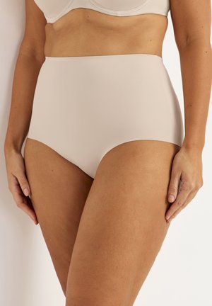 Vrouw die naadloos, hooggetailleerd nude ondergoed en bijpassende beha draagt, staande met handen langs haar lichaam tegen een witte achtergrond.