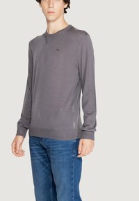 Maglione lavorato a maglia grigio a maniche lunghe con scollatura rotonda e spacchi laterali, abbinato a jeans in denim blu. Presenta un logo sottile vicino al petto.