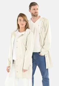 Chaquetas impermeables de color beige claro con capuchas, cierres a presión y bolsillos laterales, llevadas sobre ropa casual, con un ajuste relajado para ambos modelos.