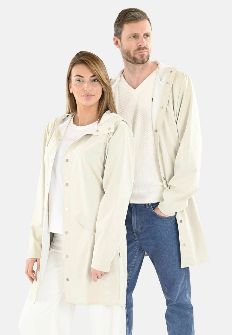 Chaquetas impermeables de color beige claro con capuchas, cierres a presión y bolsillos laterales, llevadas sobre ropa casual, con un ajuste relajado para ambos modelos.