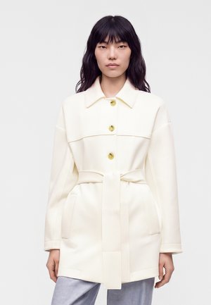 Jeune femme aux longs cheveux foncés portant un manteau ceinturé couleur crème avec de gros boutons et un pantalon gris clair sur un fond uni.