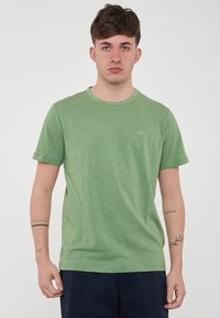 T-shirt verde a maniche corte realizzato in morbido cotone con collo a giro. Presenta un piccolo logo bianco sul petto. Il tessuto ha una finitura liscia e leggermente strutturata.