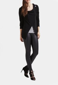 Svart topp i omlottstil av mjukt material, lager-på-lager över en ljusgrå skjorta; kombinerad med slanka svarta leggings och klackade sandaletter med snörning.