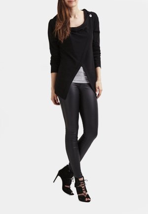 Czarny top w stylu kopertowym z miękkiego materiału, noszony na jasnoszarej koszuli; zestawiony z eleganckimi czarnymi legginsami i sandałami na obcasie z wiązaniami.