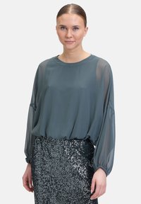 Blusa a maniche lunghe, di colore teal, in un tessuto leggero con un taglio morbido, abbinata a una gonna nera scintillante con paillettes. La texture liscia contrasta con le paillettes.