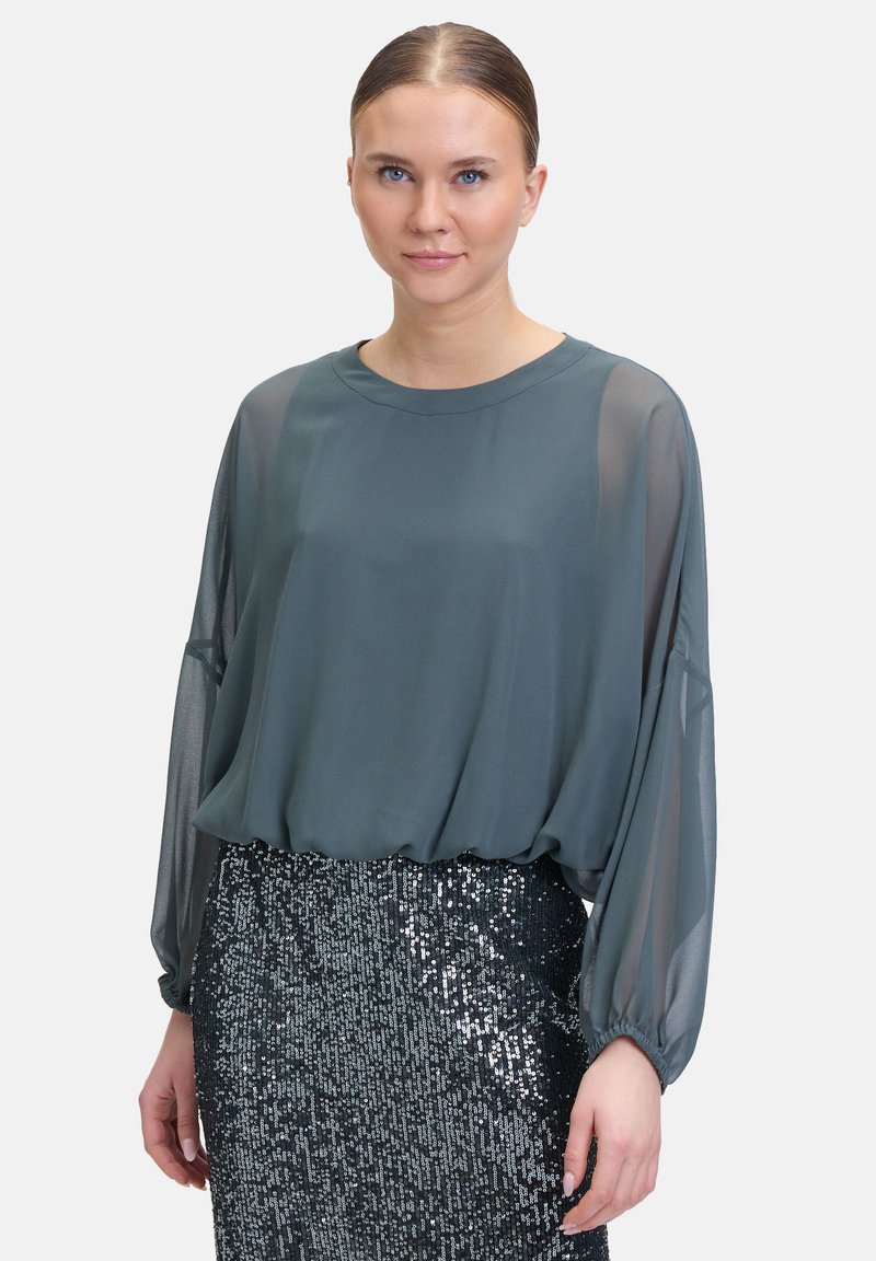 Blusa a maniche lunghe, di colore teal, in un tessuto leggero con un taglio morbido, abbinata a una gonna nera scintillante con paillettes. La texture liscia contrasta con le paillettes.