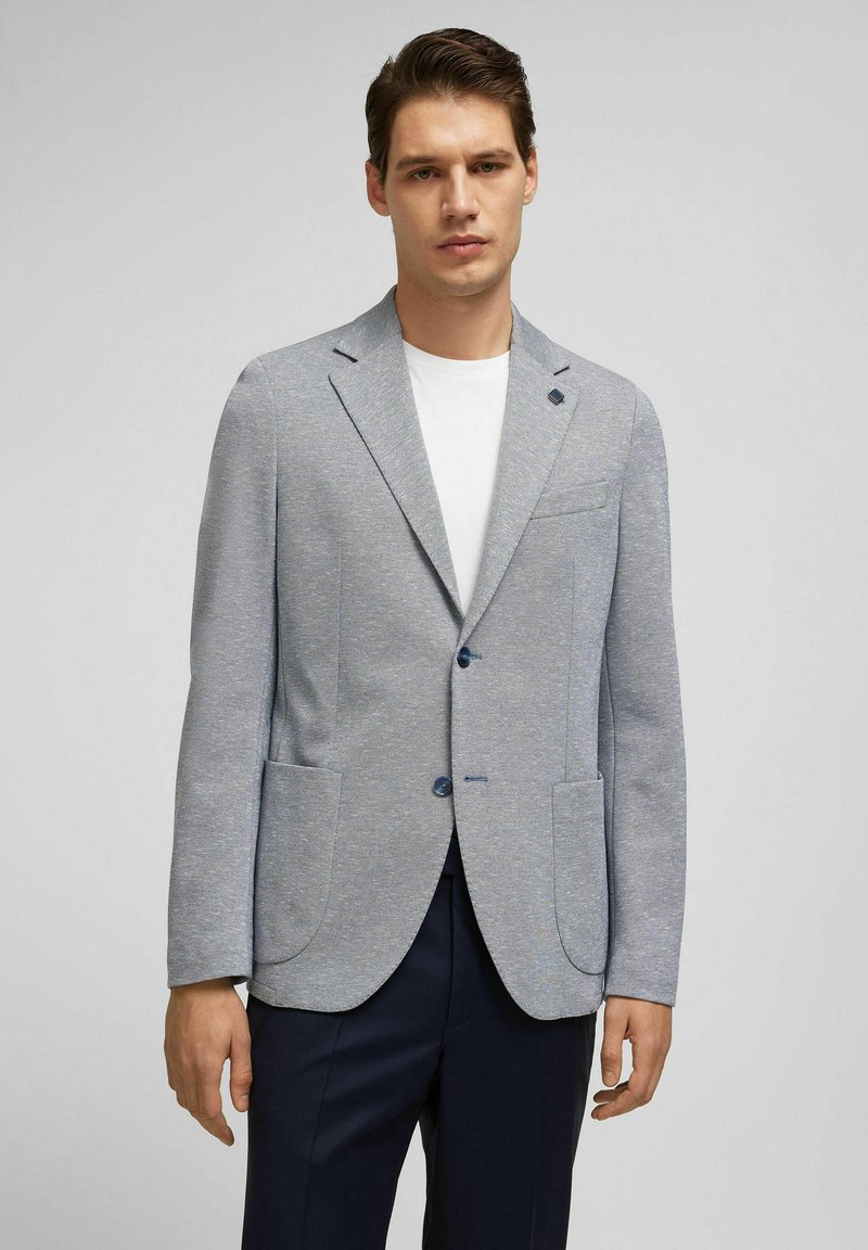 Hellgrauer, strukturierter Blazer mit Reverskragen, zwei aufgesetzten Taschen auf der Vorderseite und blauen Knopfakzenten. Stylisch über einem weißen T-Shirt getragen.