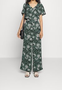Jumpsuit floreale in tessuto verde, con maniche corte, gambe larghe e scollo a V. Decorato con motivi floreali e fogliari bianchi.