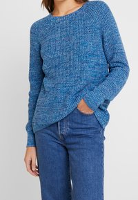 Blå stickad tröja med rund halsringning, strukturerat mönster och långa ärmar, kombinerad med denimjeans. Har en avslappnad passform.