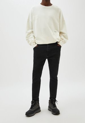 Light beige long-sleeve sweatshirt with a patch pocket, paired with black slim-fit jeans and black sporty boots. Minimalist design, no patterns.