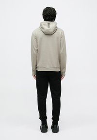 Világos bézs kapucnis pulóver húzózsinórral, amelyen függőleges csíkminta látható a hátoldalon; fekete melegítőnadrággal és fekete cipővel párosítva.