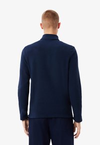 Giacca in pile blu navy con colletto, maniche lunghe e una texture morbida. Design minimalista, senza loghi o motivi visibili.