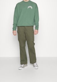 Zelená mikina s dlouhým rukávem a logem "CHAMPION" je spárována s olivovými cargo kalhotami s bočními kapsami a černobílými teniskami s potiskem.