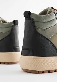 Bottes montantes en vert olive et noir. Tissu texturé et semelle en caoutchouc avec des crampons proéminents. Surpiqûres contrastées et languette de tirage.