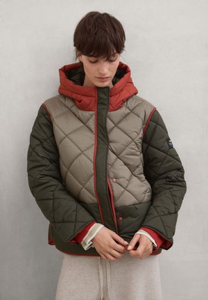Persona con el pelo corto que lleva una chaqueta acolchada en verde, beige y rojo, abrochando la cremallera frontal mientras mira hacia abajo.
