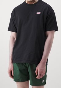 Svart bomullströja med rund hals och korta ärmar, med en liten broderad röd och grå sneaker på bröstet, tillsammans med gröna shorts.