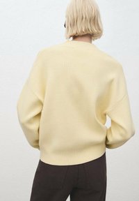 Suéter amarillo con canalé de corte relajado, hombros caídos y cuello redondo, combinado con pantalones marrón oscuro, visto desde la parte de atrás.