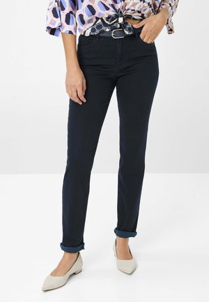 Dunkelblaue Slim-Fit-Jeans mit aufgerollten Säumen, aus glattem Stoff gefertigt. Kombiniert mit hellen, spitzen Flats und einer gemusterten Bluse.