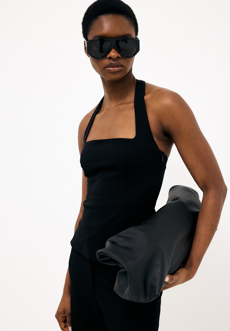 Ženska, ki nosi velike črne sončna očala in črno brezrokavnik, pod roko drži velik zvit predmet iz črne kože.