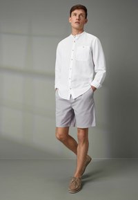Mann in weißem, knopflosem Hemd mit Kragen, hellgrauen Shorts und beigen Bootsschuhen vor grauem Hintergrund.