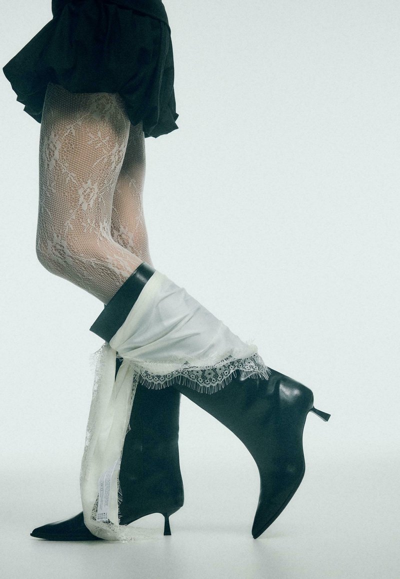 Bottes noires hauteur genou avec un bout pointu et un talon stiletto, partiellement recouvertes d'un tissu blanc orné de dentelle. Collants texturés et à motifs en dessous.