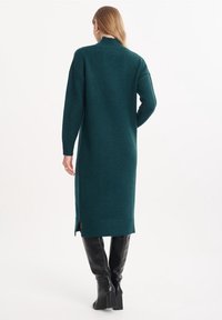 Teal stickad klänning med hög hals, långa ärmar, sidoklyftor och ribbade muddar, tillsammans med svarta knähöga stövlar med blockklack.