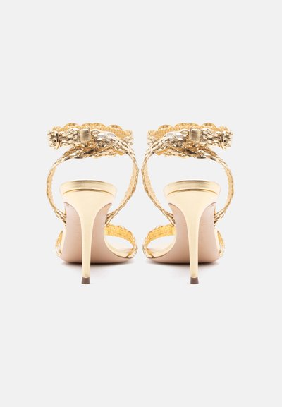 Ζευγάρι από χρυσά σανδάλια με υψηλό τακούνι, πλεγμένα λουριά και πόρπες στον αστράγαλο, σε θέα από πίσω σε λευκό φόντο.