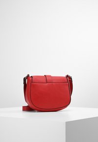 Rød læder crossbody taske med rund bund og justerbar rem, placeret på hvidt pedestalskilt mod en ensfarvet baggrund.