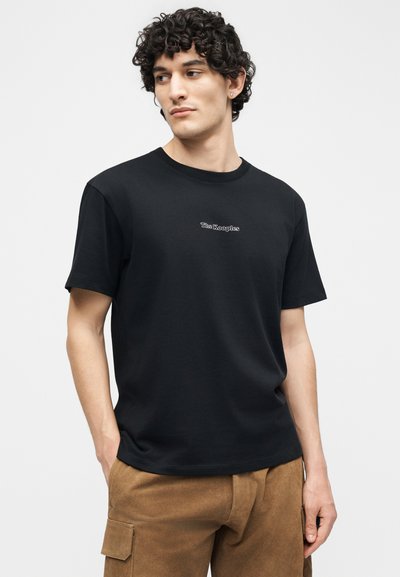 Νεαρός με σγουρά μαλλιά που φοράει μαύρο μπλουζάκι «The Kooples» και καφέ παντελόνι cargo, με τα χέρια στις τσέπες, κοιτάζει αριστερά.