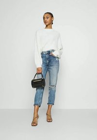 Eine Frau trägt einen weißen Strickpullover, zerrissene blaue Jeans, Riemchenschuhe und hält eine kleine schwarze Handtasche, während sie vor einem schlichten hellen Hintergrund steht.
