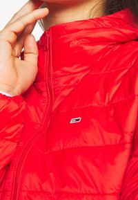 La mano di una persona che aggiusta il colletto di un giubbotto trapuntato rosso brillante, con una cerniera visibile e un piccolo logo rettangolare sul petto.