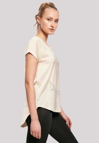 Helles beige T-Shirt mit kurzen Ärmeln, V-Ausschnitt und lockerer Passform; verfügt über ein schwarzes Pfeil-Design auf der Vorderseite und einen abgerundeten Saum.