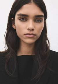 Zbliżenie portretu osoby o ciemnych, prostych, do ramion włosach, ubranej w czarną bluzkę, na jasnym tle. Neutralny wyraz twarzy.