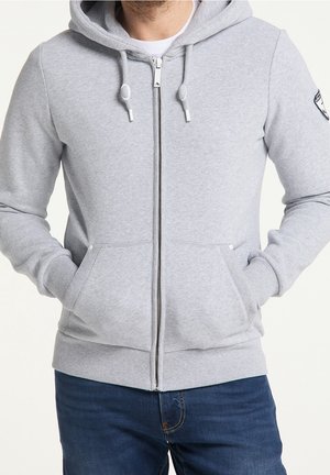 Homme portant un sweat à capuche zippé gris clair avec cordons de serrage et poches frontales, associé à un jean bleu foncé, mains dans les poches.