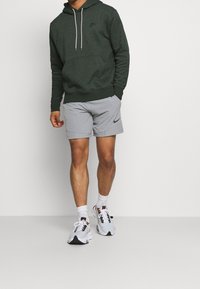 Tmavě zelená mikina s nastavitelnými šňůrkami, přední kapsou a černým logem; šedé šortky; bílé a šedé tenisky s tmavými akcenty.