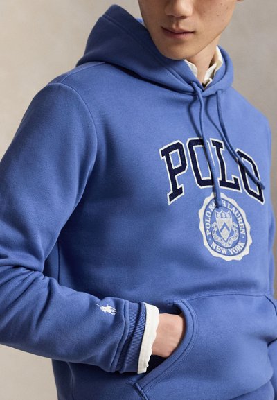 Άτομο που φοράει μπλε φούτερ Polo Ralph Lauren με λογότυπο και έμβλημα στο στήθος, χέρι μερικώς στην μπροστινή τσέπη και λευκή μανσέτα πουκάμισου ορατή.