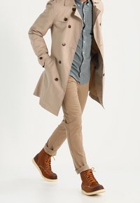 Beige trenchcoat met donkere knopen, gelaagd over een geruite shirt, gecombineerd met tan broek en een paar bruine veterschoenen met oranje veters.