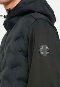 Sort polstret jakke med hette, med et glatt stoff og en rund logopatch på venstre erme, som viser en matt tekstur.