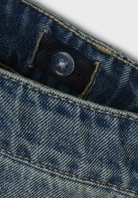Detalj traperica prikazuje plavu tkaninu s horizontalnom rebrastom teksturom, prozračnim plastičnim dugmetom i kontrastnim žutim šavovima.